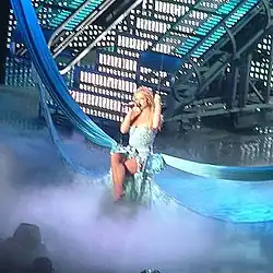 Distant image of a blond woman. She is sitting on a swing hanging from two pieces of fabric. Smoke surrounds her. She is wearing a dress and has her legs crossed. She is holding a microphone and grabbing the swing.