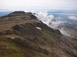 "A smattering of buildings situated on a flat-topped mountain with fog rolling off one side."