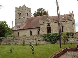 A stone church with red tiled roofs seen from the south. Extending from the chancel on the right is a battlemented chapel and, to the left, the tower is also battlemented