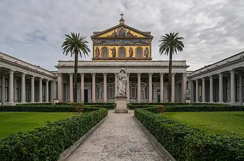 A courtyard with palm trees and a greater-than-lifesized statue of Saint Paul holding a sword in front of the colossal portico of the basilica and a large mural covering the upper facade