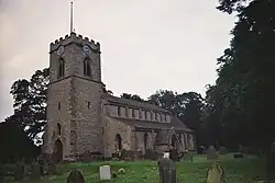 A small church with a square Ashlar tower, in the Early English style. The tower is to the left, the nave to the right, and we are looking somewhat obliquely at it. The church is ringed by medium size trees, and a large Yew dominates the right of the picture, in the middle distance. A handful of mismatched gravestones dot the grassy nearground of the picture.