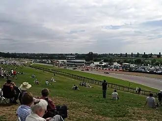 View of the track stretching away to the left from an elevated grassy bank with spectators, showing circuit buildings of far side of track with lakes beyond against a dappled sky