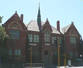 A photograph of Carlton Gardens Primary School, taken during the day. A two-floor red brick building that has many windows and a sign above its front door that reads "No. 2605".