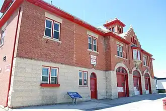 A two-storey concrete and red brick fire hall. The front of the hall has three large doors for vehicles, one regular door, and two windows.