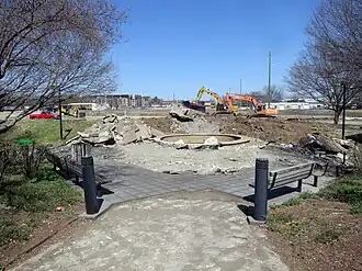 Construction equipment has begun to dig up concrete and level the earth beyond an area with a concrete fountain and wooden benches.