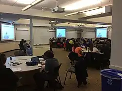 Phot of the MIT technology-enhanced active learning (TEAL) classroom with multiple screens, white boards data projectors and round tables where students work together.