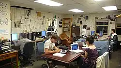 Several young men and women sit in office chairs working at computers around a room where the walls are covered in printed pages. A central wood table and bookcases are featured.