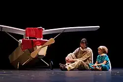 A prop plane with several pieces of cloth on top of it sits to the right of a man in pilot gear and a young child in pajamas.