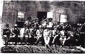 A black-and-white photograph of a group of men standing and sitting in front of a war damaged building. The caption at the bottom lists the names of those present