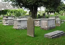 Three-quarter view of several large stone tombs and some gravestones in a grassed churchyard, with trees visible in the background.
