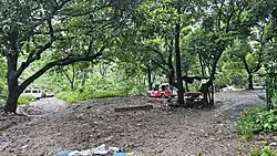 A three-way intersection of dirt roads with significant tree cover and some parked motor vehicles.