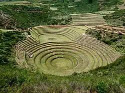 Photo of a large pit in the mountain seen from above. A round pit is surrounded by 7 levels of terraces. The terrace extends onto a flat area and there are further 6 levels of terraces around. A series of 9 terraces rises further in the background. The terraces are covered with short grass. The surrounded area is covered with darker wild bushes.