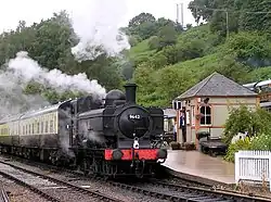 A black pannier tank locomotive is waiting at a platform with a train of two cream and brown passenger carriages. In the background is a green wooded hill, and a small wooden station building with a slate roof. The station master is sheltering from the rain in the doorway of the building. Smoke and steam are coming from the locomotive as it prepares to move off.