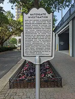 Photo of a grey historical marker titled "Watergate Investigation" on the sidewalk beside a small urban street with a parking garage door visible on the right behind the sign. The marker reads: "Mark Felt, second in command at the FBI, met Washington Post reporter Bob Woodward here in this parking garage to discuss the Watergate scandal. Felt provided Woodward information that expose the Nixon Administration's obstruction of the FBI's Watergate investigation. He chose this garage as an anonymous secure location. They met at this garage six times between October 1972 and November 1973. The Watergate scandal resulted in President Nixon's resignation in 1974. Woodward's managing editor, Howard Simons, gave Felt the code name 'Deep Throat'. Woodward's promise not to reveal his source was kept until Felt announced his role as Deep Throat in 2005. Erected in 2008 by Arlington County, Virginia."