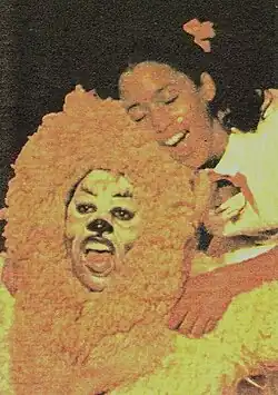 Young man in lion costume kneels in front of young girl with pigtails, both singing