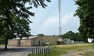 A one-story gray metal building with a W L I W logo next to the base of a tall broadcasting tower
