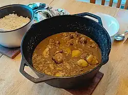 A cast-iron pot of chicken and potato xacuti, a warm yellow-brown curry, sits on a table. There is a small pot of rice visible in the top right of the frame.