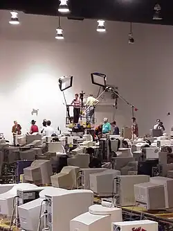 Television crews and filming equipment on a platform above tables with rows of CRT monitors in a convention hall